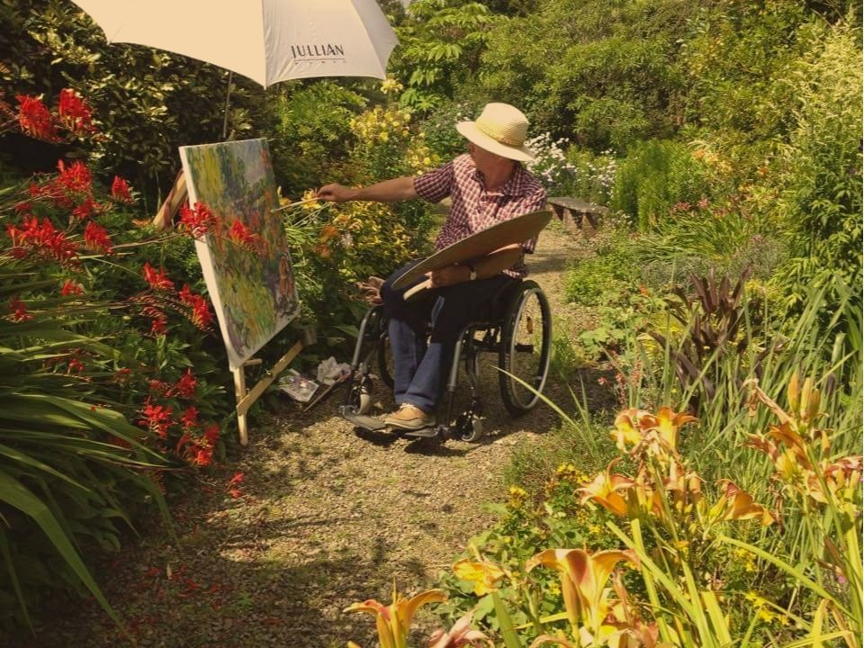 Person in a wheelchair painting in a garden using The Nadeau Easel because of it's accessibility features.